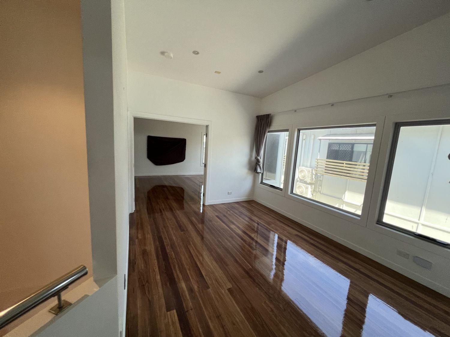 Empty room with shiny wooden floors, large windows, and an adjacent room with a mounted television.
