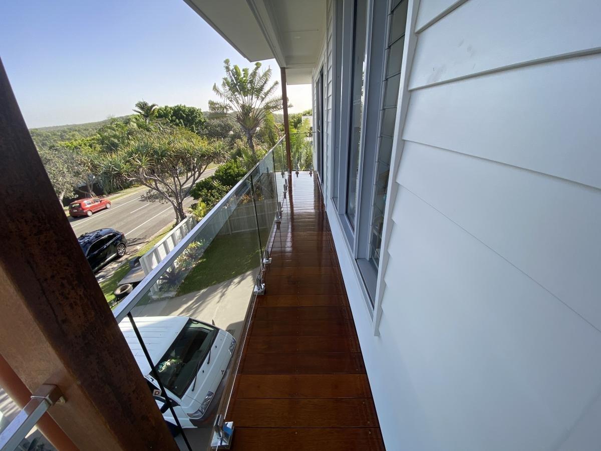 Balcony with wooden flooring and glass railing overlooking a leafy street.